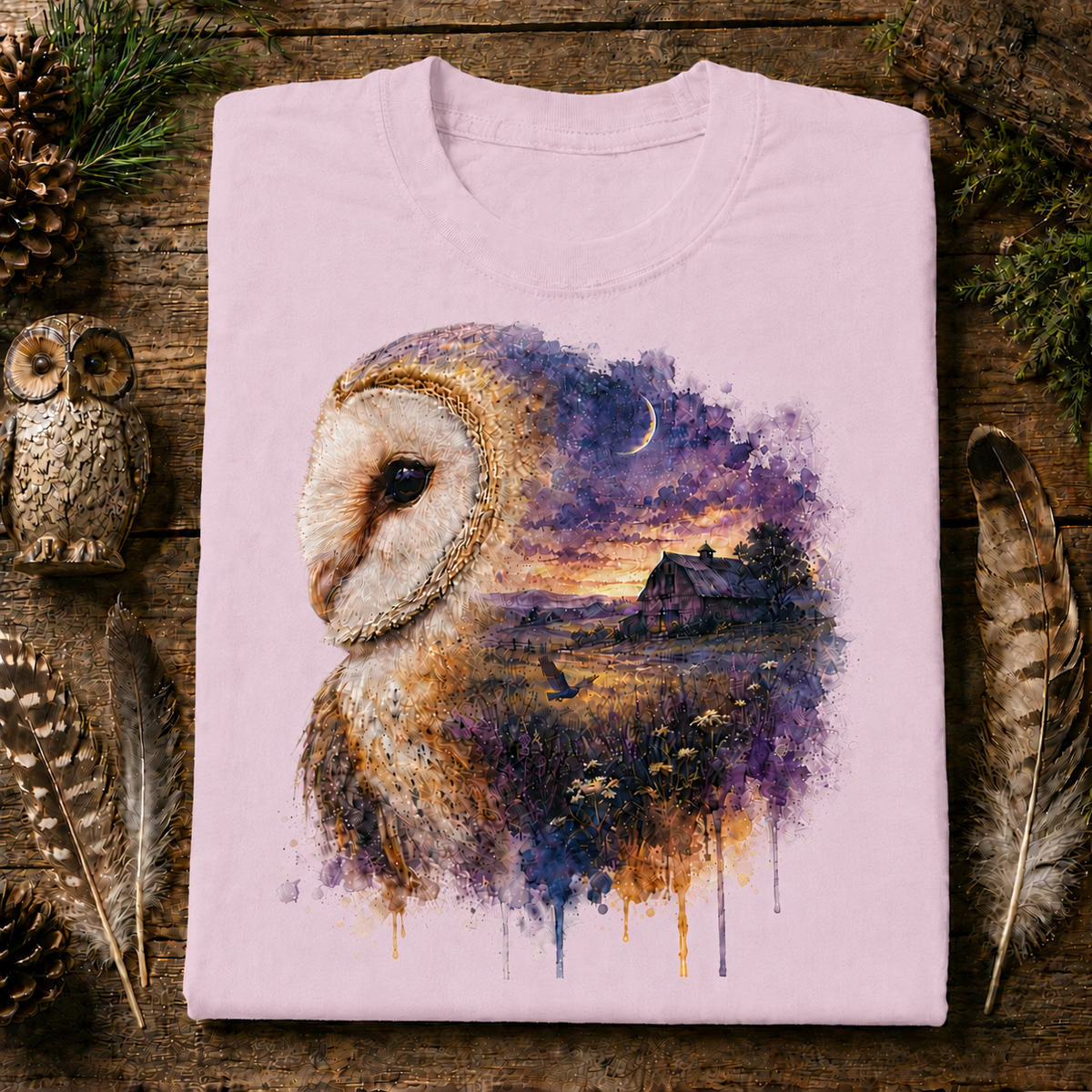 A folded blossom colored t-shirt with a detailed watercolor graphic of an owl and a rustic barn landscape, displayed on a rustic wooden background with decorative feathers and pinecones.