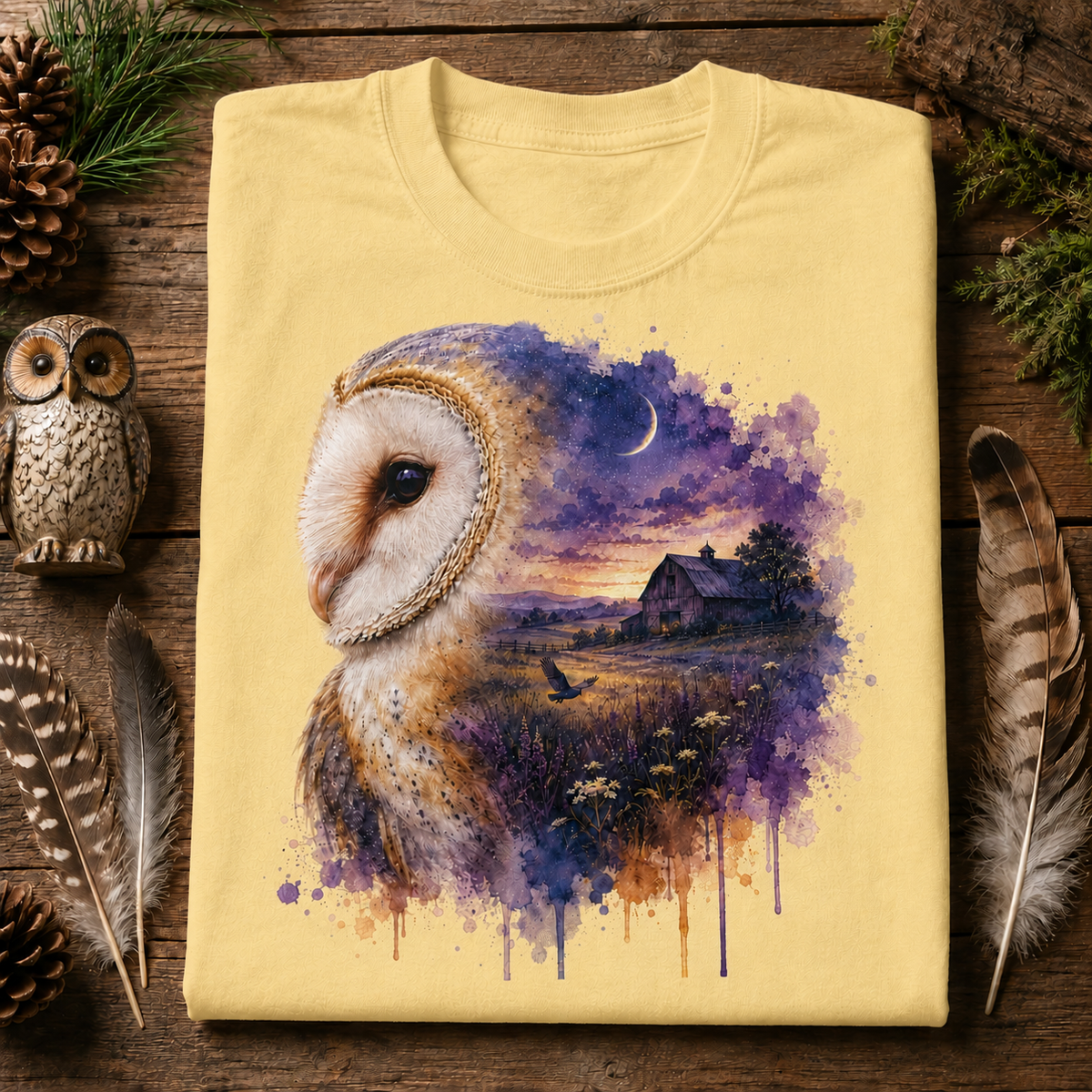 A folded butter colored T-shirt laid flat on a rustic wooden surface, featuring a large printed artwork of a barn owl in profile with a purple and blue watercolor landscape, barn, moon, and bird. Decorative feathers, pinecones, and a small owl figurine surround the shirt.