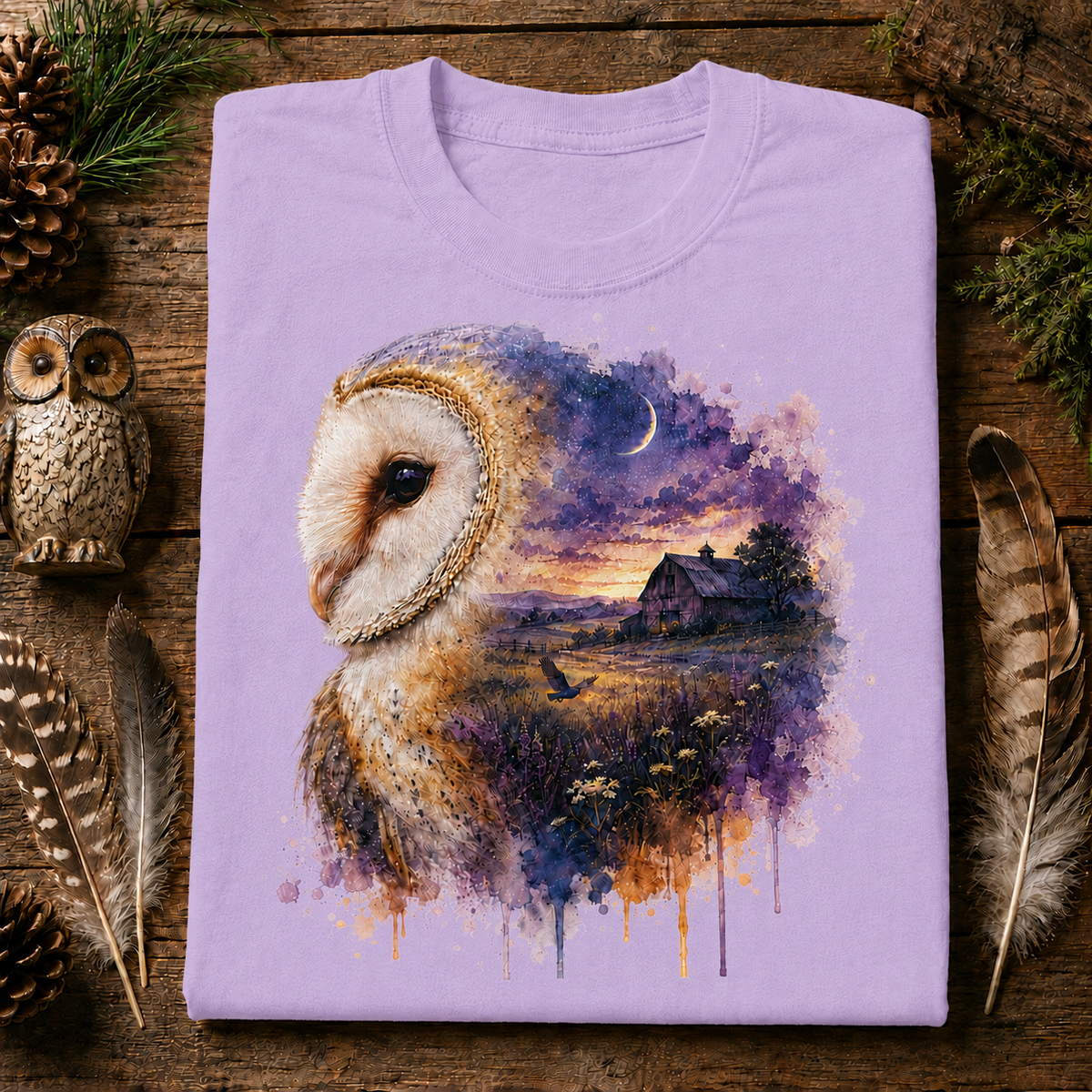 A folded orchis colored crewneck t-shirt with a detailed graphic print of a barn owl and a watercolor landscape, displayed on a rustic wooden background with decorative feathers, pinecones, and a wooden owl figurine.