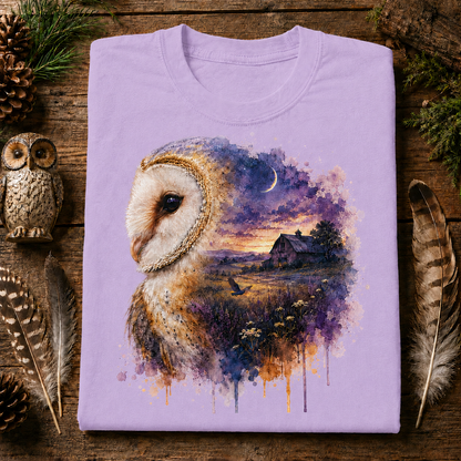 A folded orchis colored crewneck t-shirt with a detailed graphic print of a barn owl and a watercolor landscape, displayed on a rustic wooden background with decorative feathers, pinecones, and a wooden owl figurine.