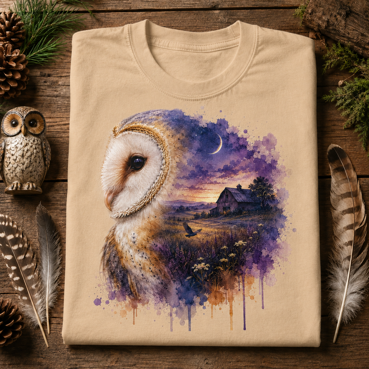 A sandstone colored crewneck t-shirt is laid flat on a rustic wooden surface, decorated with pine needles, pinecones, feathers, and a small owl figurine. The t-shirt features a detailed graphic print of a barn owl next to a purple and blue watercolor landscape of a barn at dusk.