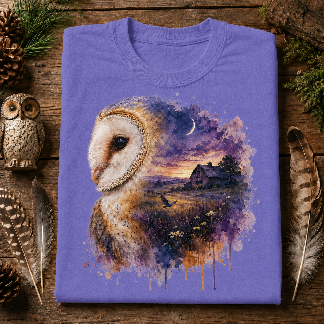A folded violet  colored crewneck t-shirt with a detailed graphic of a barn owl and a watercolor landscape, displayed on a rustic wooden background with decorative feathers, pinecones, and a small owl figurine.