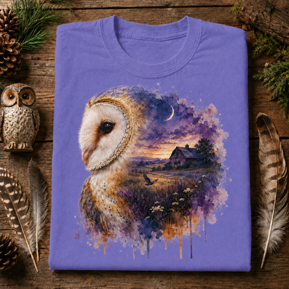 A folded violet  colored crewneck t-shirt with a detailed graphic of a barn owl and a watercolor landscape, displayed on a rustic wooden background with decorative feathers, pinecones, and a small owl figurine.