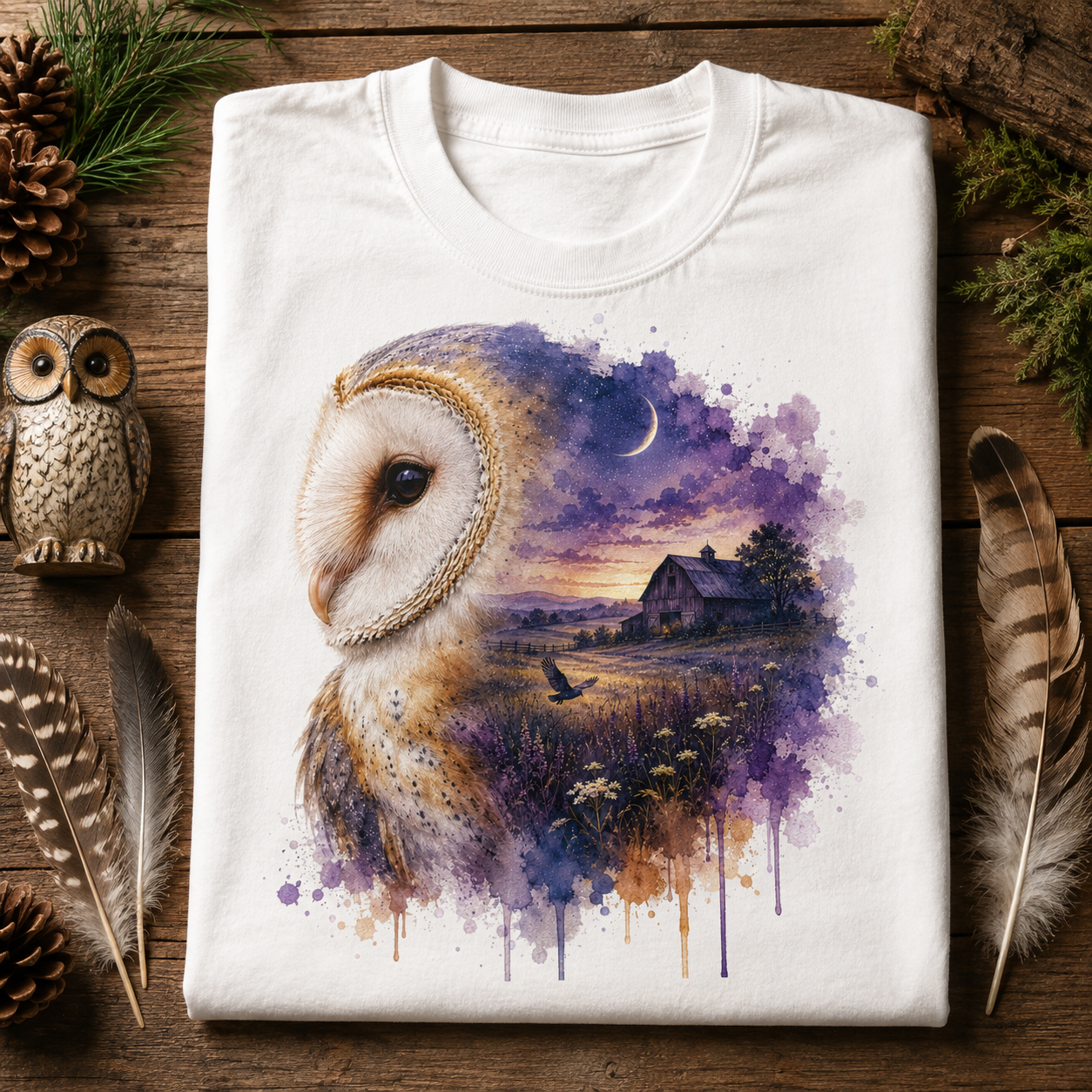 A folded white crewneck t-shirt displayed on a rustic wooden surface, decorated with pinecones, feathers, and a small owl figurine. The t-shirt features a detailed graphic print of a barn owl in profile, blending into a watercolor landscape of a farm field under a purple and blue night sky with a crescent moon.