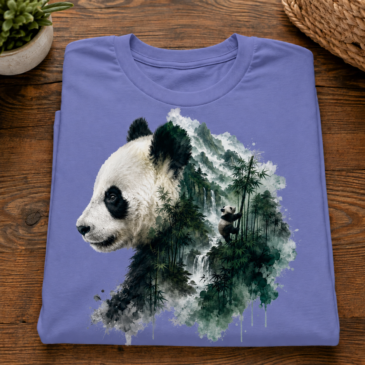 A folded violet colored t-shirt with a graphic of a panda overlaid with a green and white bamboo forest, displayed on a wooden table.