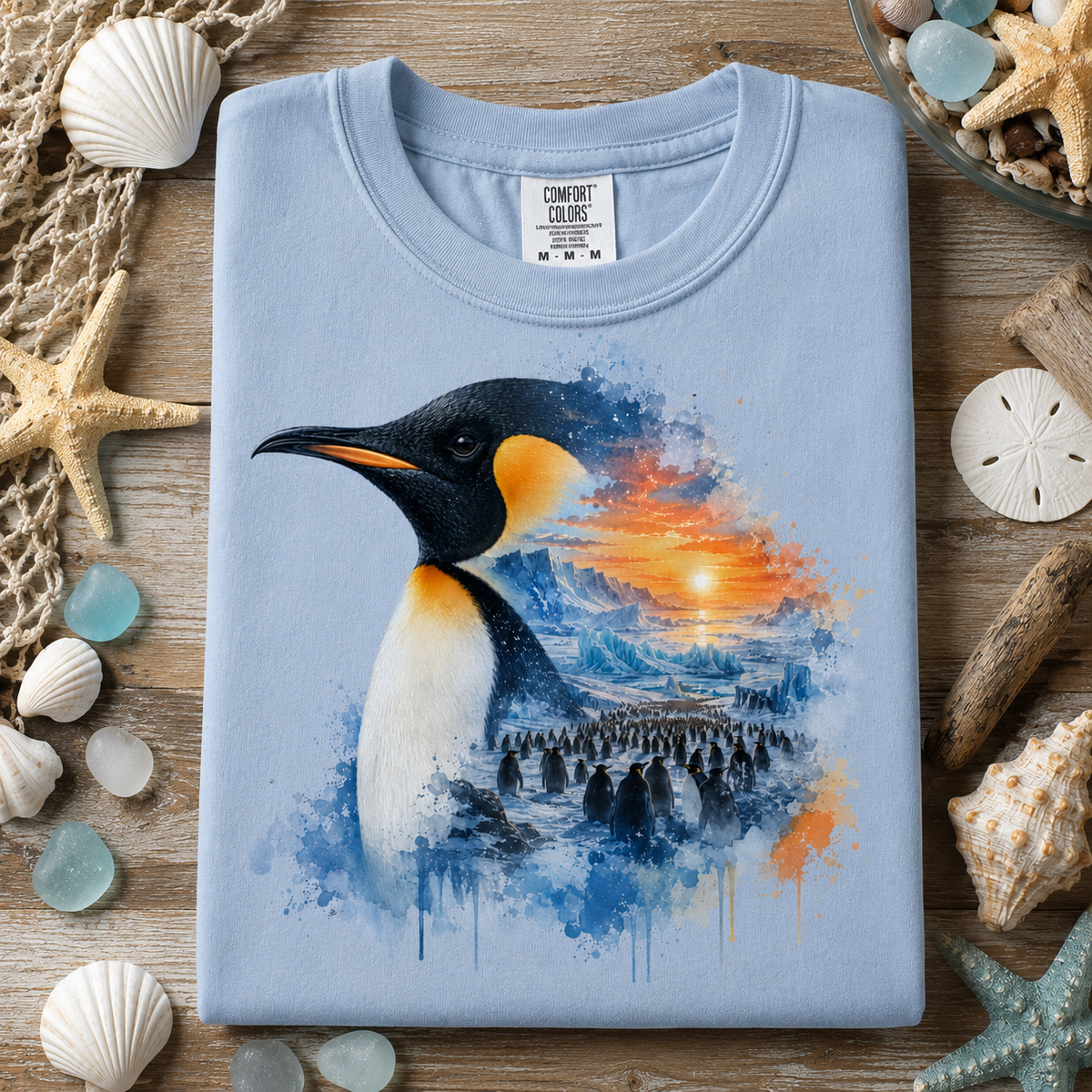 A folded light blue (hydrangea) t-shirt with a watercolor-style penguin graphic, styled in a flat-lay on a wooden surface surrounded by seashells, starfish, and driftwood.
