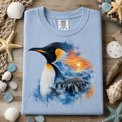 A folded light blue (hydrangea) t-shirt with a watercolor-style penguin graphic, styled in a flat-lay on a wooden surface surrounded by seashells, starfish, and driftwood.