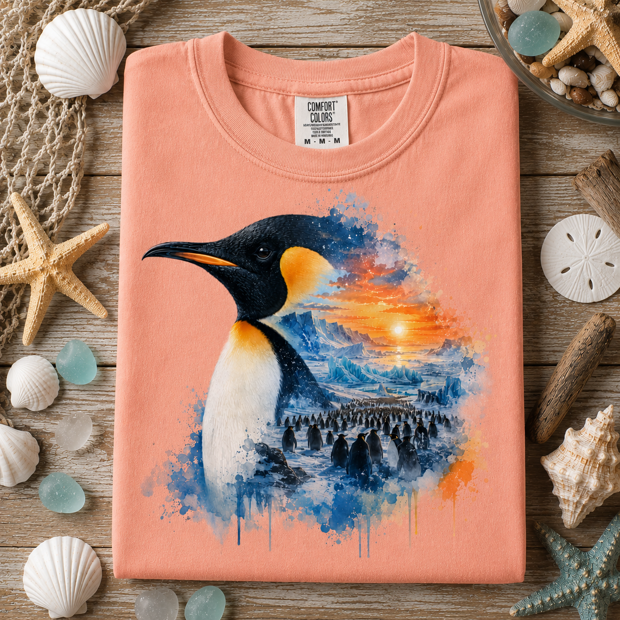 A folded peach-colored Comfort Colors t-shirt with a vibrant penguin graphic, styled in a flat-lay on a rustic wooden surface surrounded by seashells, starfish, and a net.
