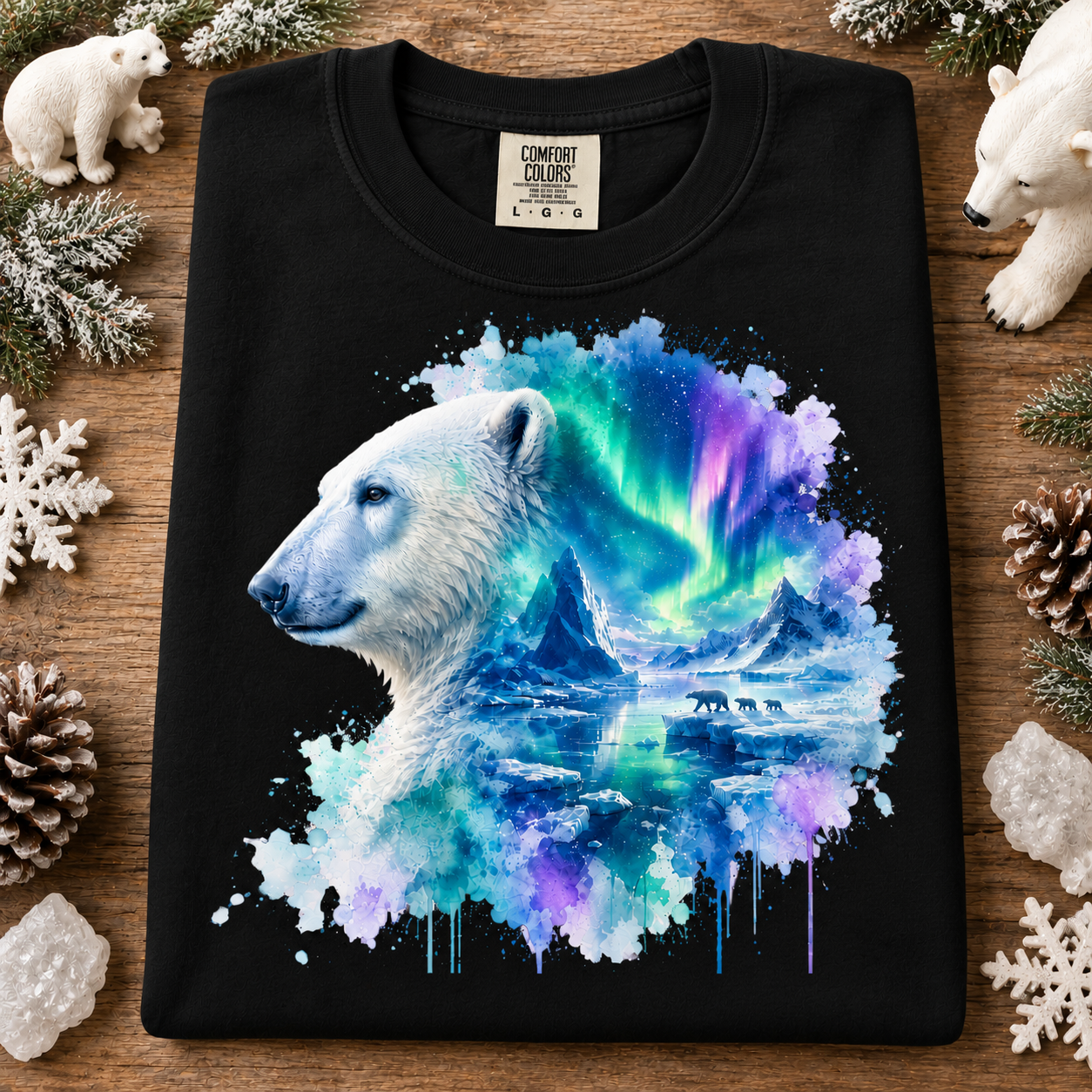 A black Comfort Colors t-shirt laid flat on a rustic wooden surface, decorated with white polar bear figurines, pinecones, and snowflakes. The shirt features a graphic print of a white polar bear profile merging into a vibrant watercolor depiction of the northern lights over icy mountains.