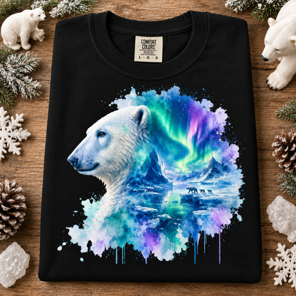 A black Comfort Colors t-shirt laid flat on a rustic wooden surface, decorated with white polar bear figurines, pinecones, and snowflakes. The shirt features a graphic print of a white polar bear profile merging into a vibrant watercolor depiction of the northern lights over icy mountains.