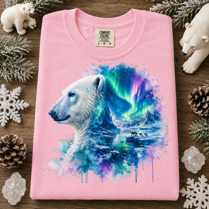 A folded blossom colored t-shirt with a colorful graphic of a polar bear and aurora borealis, styled on a wooden surface with polar bear figurines, pinecones, and snowflakes.