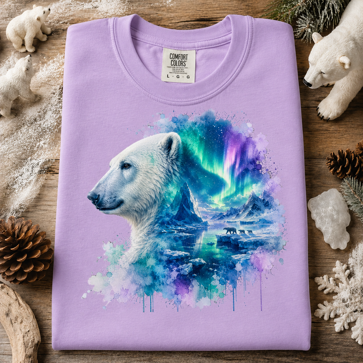 A folded orchid colored crewneck t-shirt with a graphic print of a polar bear and an aurora borealis landscape. The shirt is displayed on a rustic wooden background surrounded by winter-themed props including pinecones, a snowflake, crystals, and plush polar bear toys.