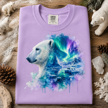 A folded orchid colored crewneck t-shirt with a graphic print of a polar bear and an aurora borealis landscape. The shirt is displayed on a rustic wooden background surrounded by winter-themed props including pinecones, a snowflake, crystals, and plush polar bear toys.