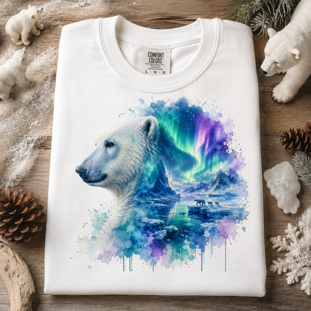 A folded white Comfort Colors t-shirt is displayed flat on a rustic wooden surface, surrounded by winter-themed props like a polar bear figurine, pine cones, and snowflakes. The t-shirt features a large graphic print of a polar bear profile merging into a vibrant watercolor scene of mountains and the aurora borealis in shades of blue, purple, and green.