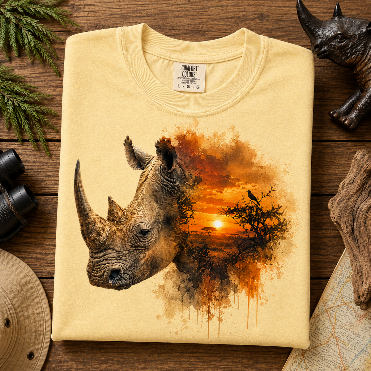 A folded butter colored Comfort Colors t-shirt with a graphic print of a rhinoceros head merging into an African savanna sunset scene, displayed on a rustic wooden background with safari-themed props.