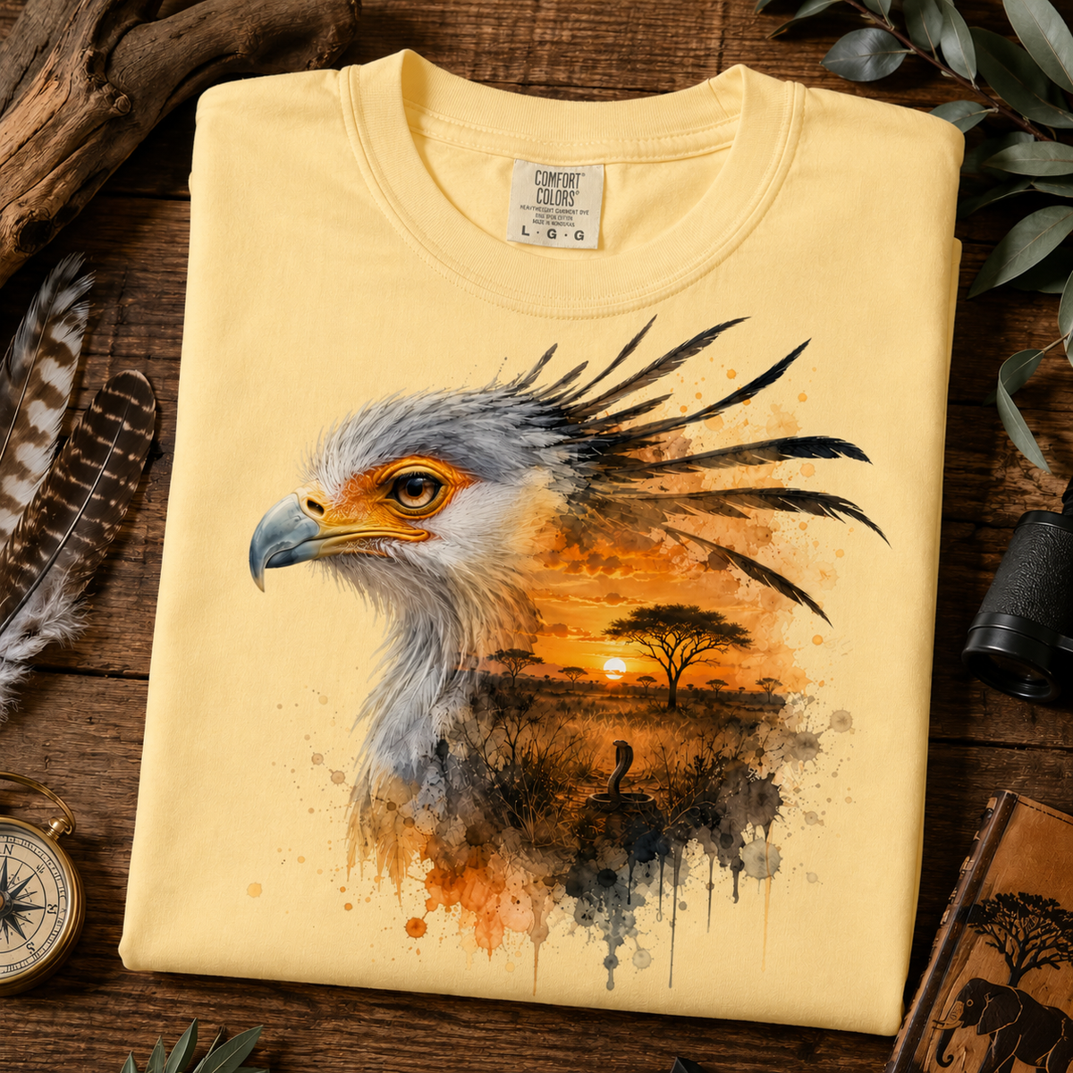 A folded butter yellow T-shirt laid flat on a rustic wooden surface, featuring a large watercolor-style print of a bird of prey head over an African savanna sunset scene. Decorative props including feathers, a compass, binoculars, and leaves surround the shirt.