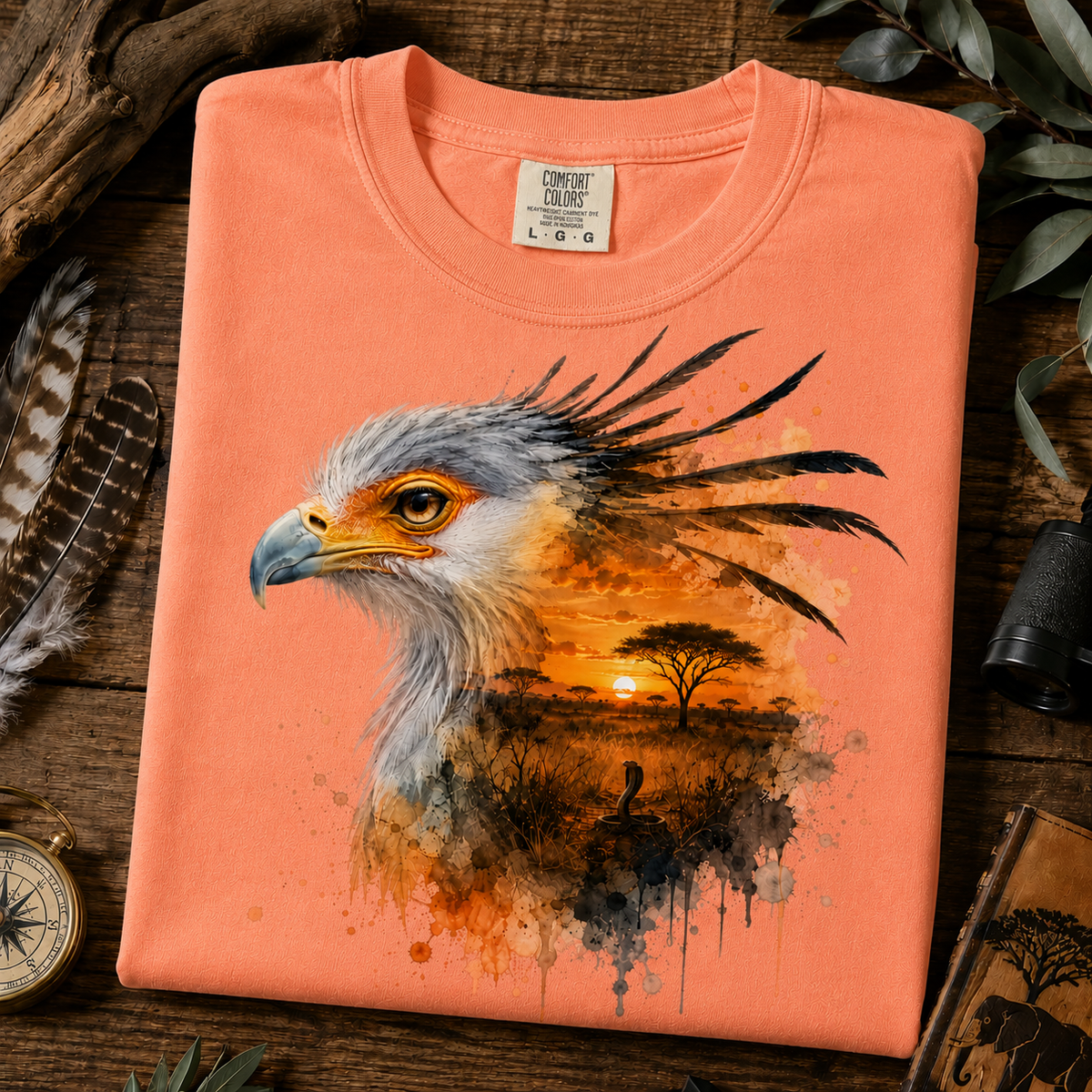 A folded melon colored crewneck t-shirt with a detailed graphic of a bird of prey over an African savanna sunset, styled on a rustic wooden background with feathers, a compass, and a telescope.