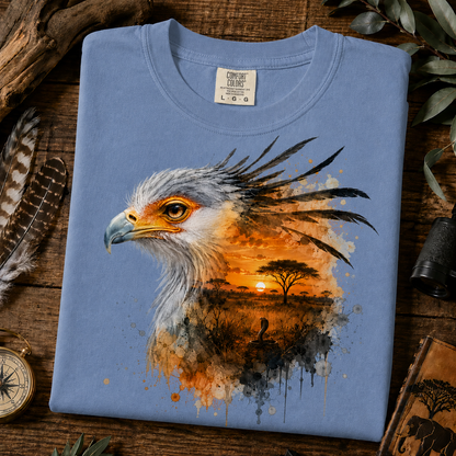 A folded washed denim colored crewneck t-shirt is displayed on a rustic wooden surface surrounded by safari-themed props, including a compass, a feather, and a binocular. The front of the shirt features a detailed graphic print of an African eagle with a watercolor sunset savanna landscape inside its feathers.