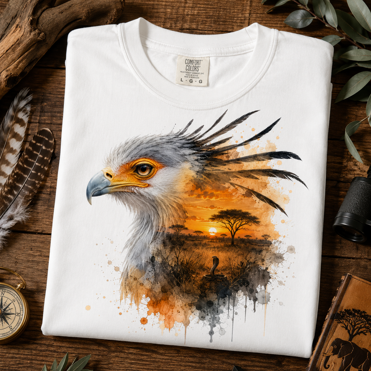 A folded white crewneck t-shirt with a detailed graphic print of a bird of prey over an African savanna sunset, styled on a rustic wooden background with feathers, a compass, and a binoculars.