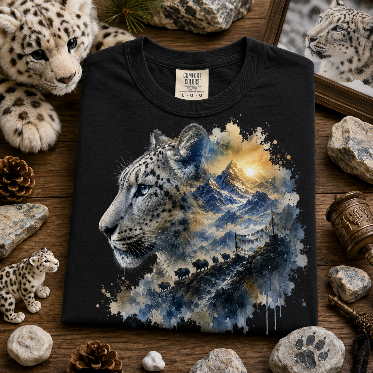 A black short-sleeve t-shirt with a detailed graphic print of a snow leopard's head merging into blue and gold mountain scenery, styled in a flat-lay on a wooden surface surrounded by rocks, pinecones, and small snow leopard figurines.