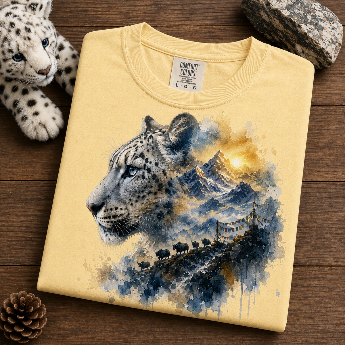 A folded butter colored T-shirt on a wooden surface, featuring a large watercolor-style print of a snow leopard with mountains, a glowing sun, and a small herd of bison. A plush toy and rocks are placed around it as styling props.