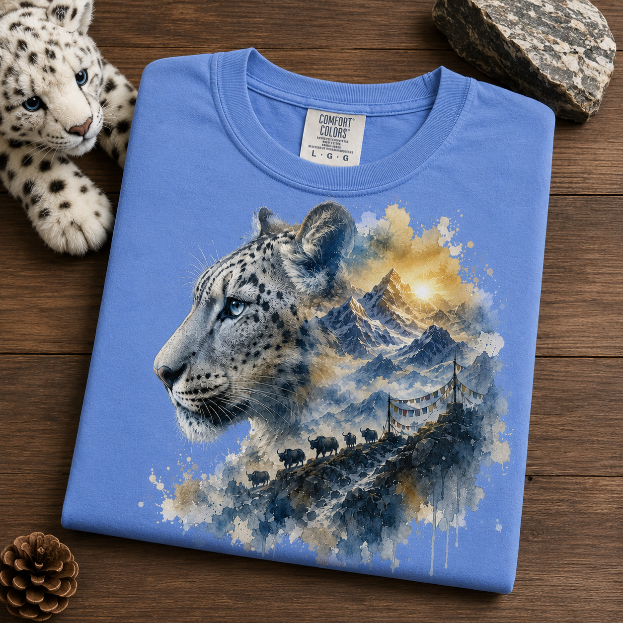 A folded mystic blue t-shirt with a detailed graphic of a snow leopard and mountain landscape, displayed on a wooden surface next to a plush toy and rocks.