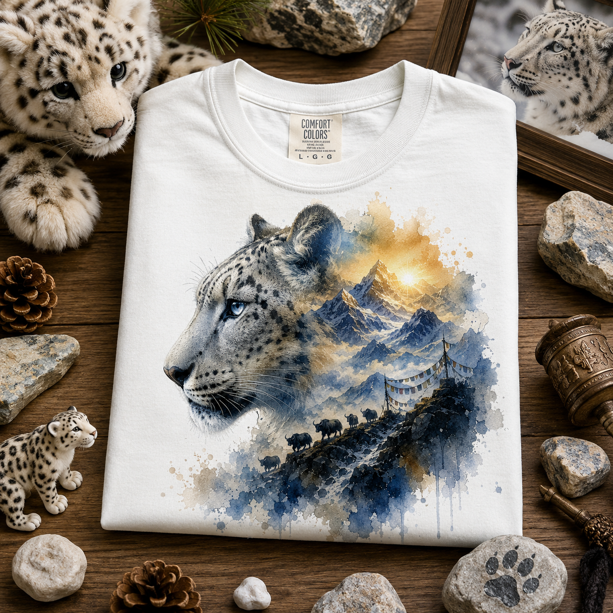 A folded white t-shirt with a detailed graphic print of a snow leopard and mountain landscape, styled in a flat-lay on a wooden surface surrounded by snow leopard toys, rocks, and pine cones.