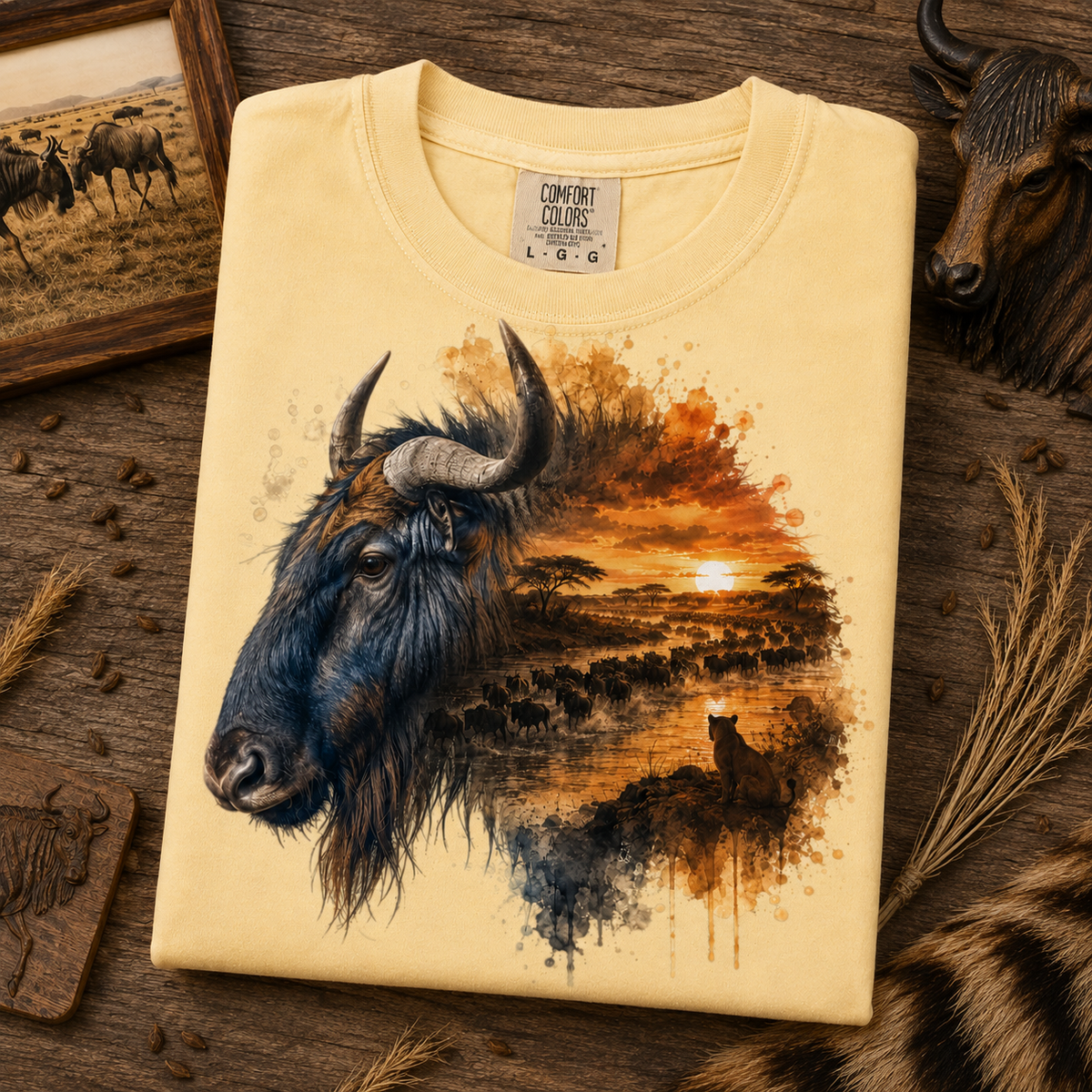 A folded butter colored Comfort Colors t-shirt featuring a graphic print of a wildebeest and a savanna sunset, styled on a rustic wooden surface with decorative animal heads and dried grass.