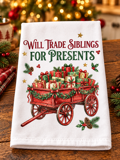 A white kitchen towel with a festive Christmas print featuring a red wagon loaded with wrapped presents and the text 'Will Trade Siblings for Presents,' displayed on a wooden table with blurred holiday decorations in the background.