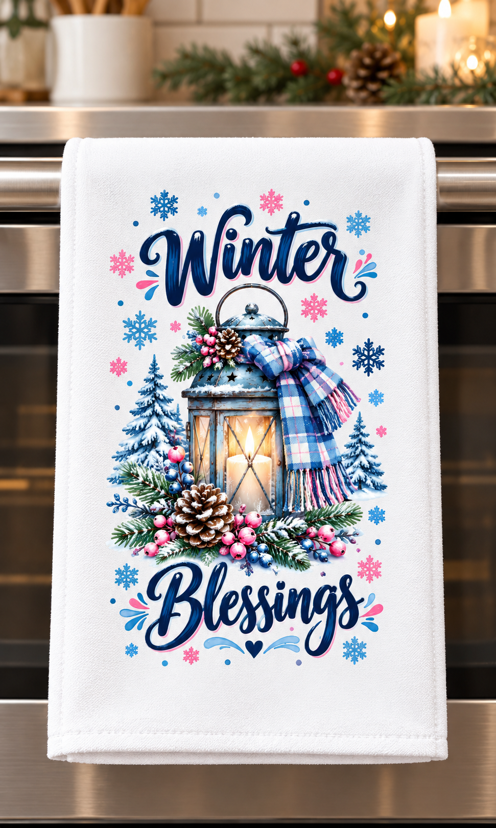 A white kitchen towel hanging on a stainless steel oven handle, featuring a winter-themed graphic with the words 'Winter Blessings' in navy script, a glowing lantern, pinecones, berries, evergreen trees, and a blue plaid scarf.