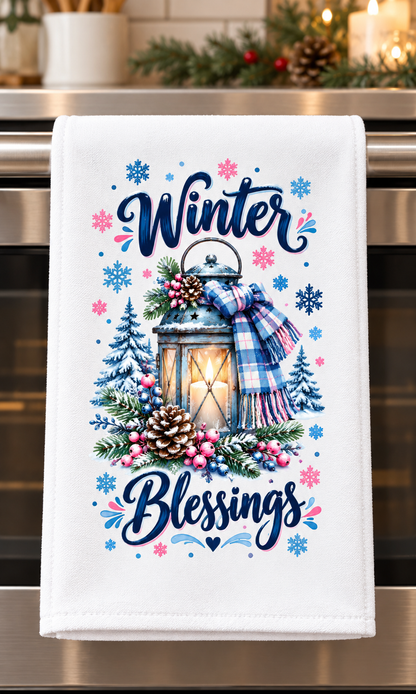 A white kitchen towel hanging on a stainless steel oven handle, featuring a winter-themed graphic with the words 'Winter Blessings' in navy script, a glowing lantern, pinecones, berries, evergreen trees, and a blue plaid scarf.