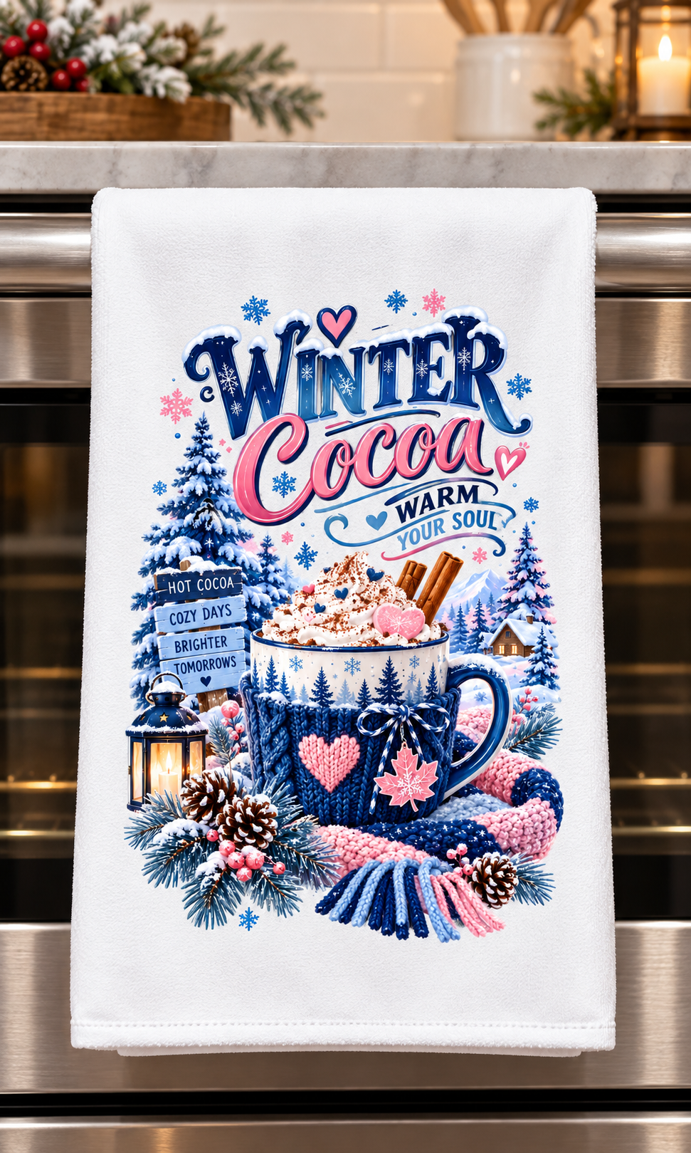 A white kitchen towel hanging on a stainless steel oven handle, featuring a colorful winter cocoa-themed graphic with a mug of hot cocoa, pine trees, snowflakes, and the words 'Winter Cocoa Warm Your Soul'.