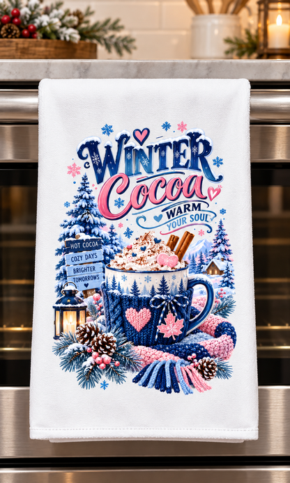 A white kitchen towel hanging on a stainless steel oven handle, featuring a colorful winter cocoa-themed graphic with a mug of hot cocoa, pine trees, snowflakes, and the words 'Winter Cocoa Warm Your Soul'.