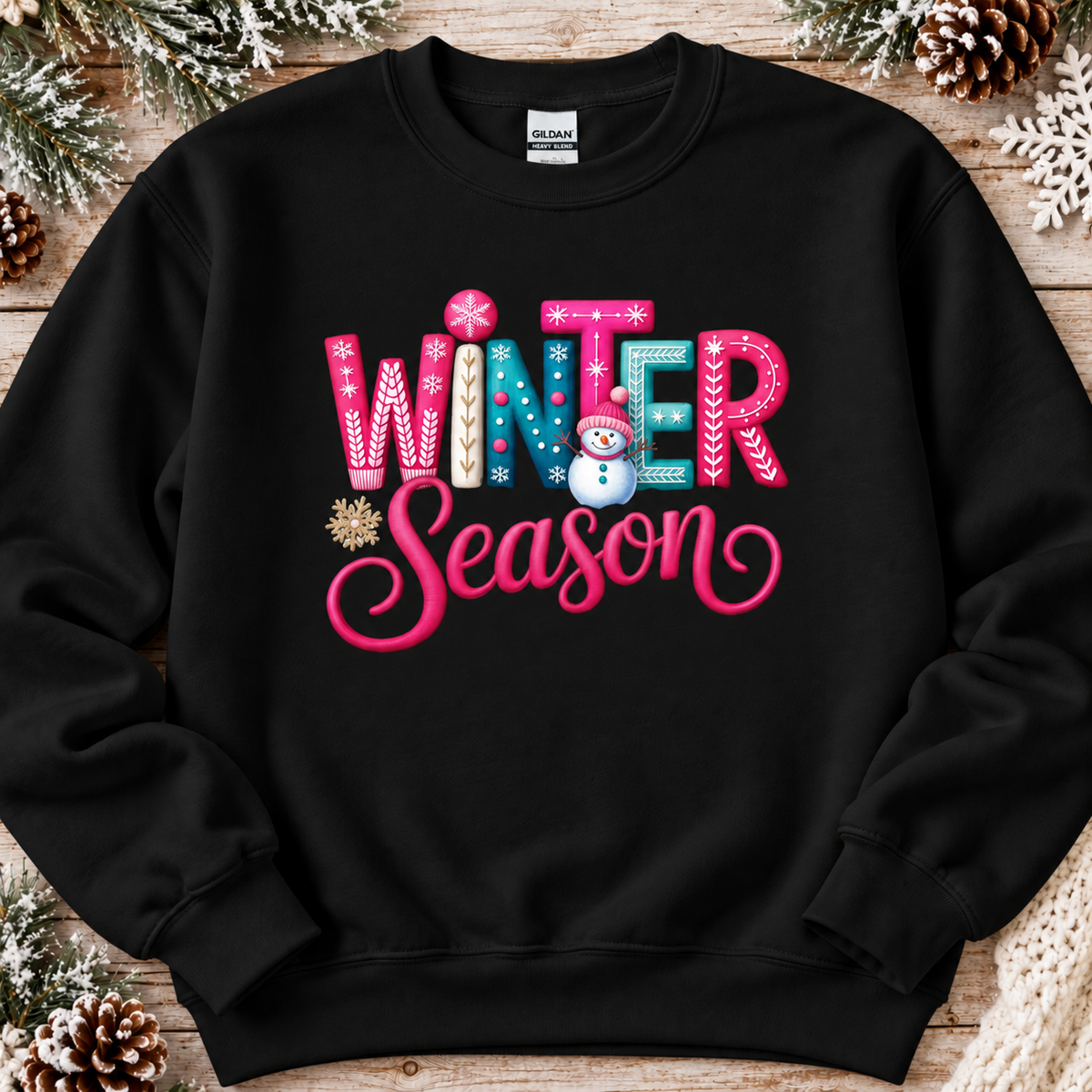 A black crewneck sweatshirt featuring a colorful graphic that reads 'WINTER Season' in pink, blue, and white decorative lettering with a small snowman and snowflake accents, displayed flat on a rustic wooden background with pinecones and snowflakes.