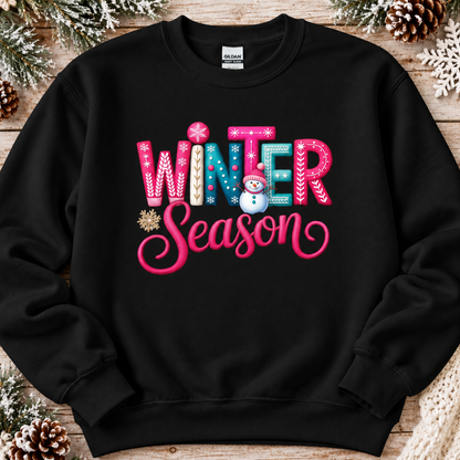 A black crewneck sweatshirt featuring a colorful graphic that reads 'WINTER Season' in pink, blue, and white decorative lettering with a small snowman and snowflake accents, displayed flat on a rustic wooden background with pinecones and snowflakes.