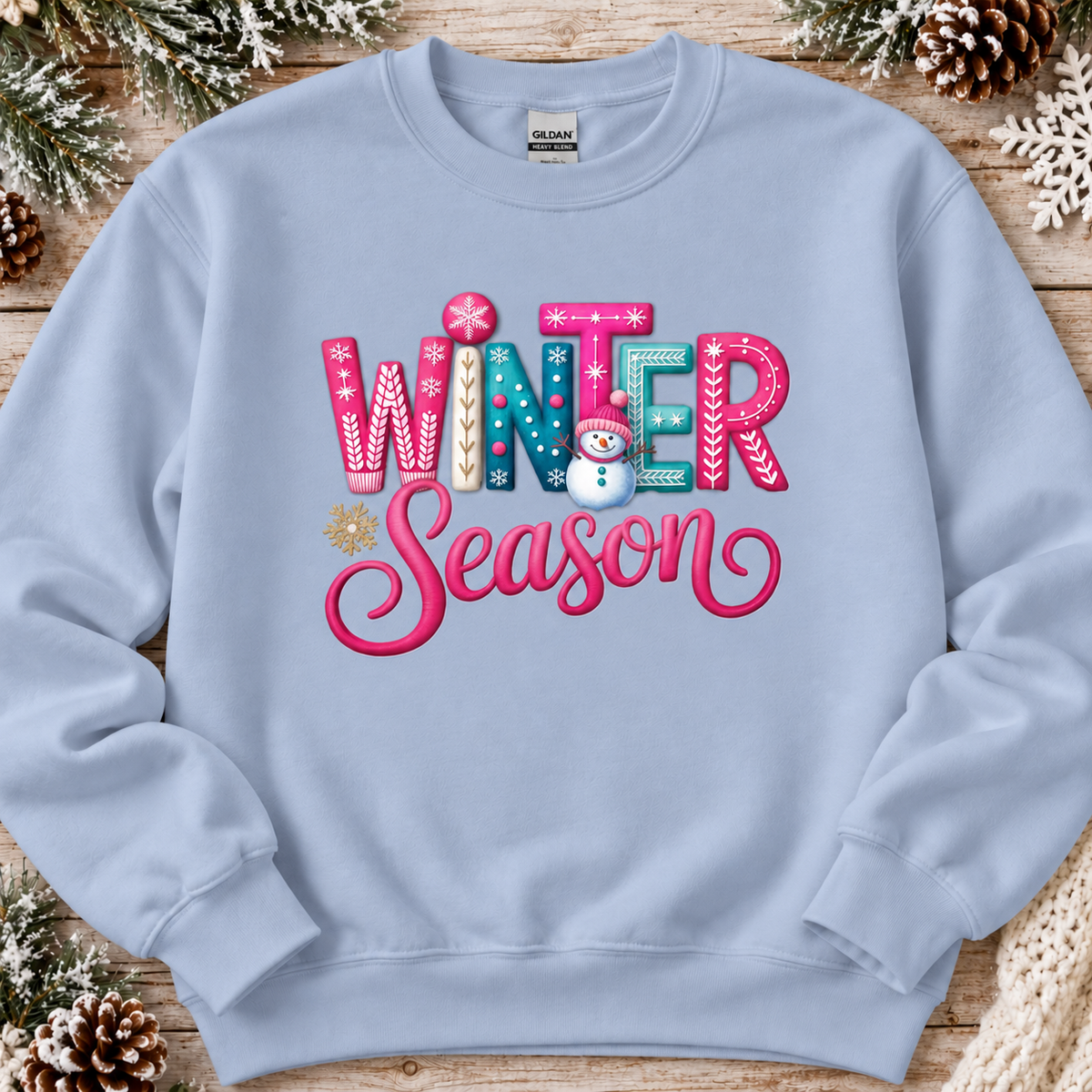 A light blue crewneck sweatshirt featuring a colorful 'Winter Season' graphic with a snowman, laid flat on a rustic wooden background decorated with pinecones and snowflakes.