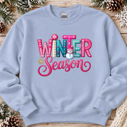 A light blue crewneck sweatshirt featuring a colorful 'Winter Season' graphic with a snowman, laid flat on a rustic wooden background decorated with pinecones and snowflakes.