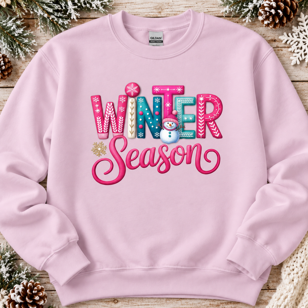 A light pink crewneck sweatshirt featuring a colorful 'WINTER Season' graphic on the front, styled flat on a rustic wood background with pine branches and snowflakes.