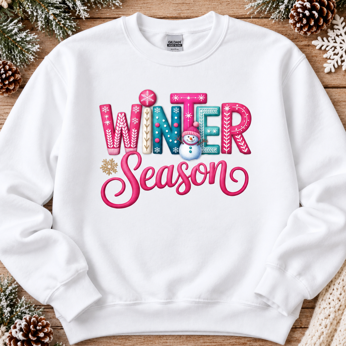 A white crewneck sweatshirt with a colorful 'Winter Season' graphic, laid flat on a rustic wooden background decorated with pine needles, pinecones, and a white snowflake.