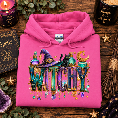 A folded azalea pink hoodie is displayed on a rustic wooden surface with Halloween-themed props, including a black candle, a spell book, a broom, a crystal ball, and gold stars. The hoodie features a large graphic on the front with the word 'WITCHY' in decorative teal and purple letters, topped with a purple witch hat, a black cat, and a crescent moon.