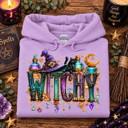 A lavender orchid purple Gildan hoodie featuring a detailed 'WITCHY' graphic print with a witch hat, potion bottles, a black cat, a crescent moon, and hanging gem charms, styled in a flat-lay on a rustic wooden table surrounded by seasonal props.