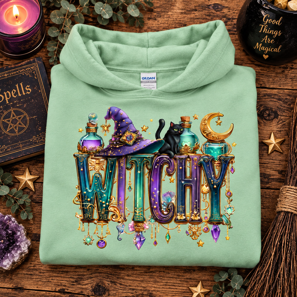 A folded mint green hoodie is displayed on a rustic wooden surface surrounded by Halloween-themed props, including a purple candle, a book of spells, a broom, and crystals. The hoodie features a large, colorful graphic on the front with the word 'WITCHY' in decorative letters, a purple witch hat, a black cat, potion bottles, and a crescent moon.