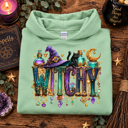 A folded mint green hoodie is displayed on a rustic wooden surface surrounded by Halloween-themed props, including a purple candle, a book of spells, a broom, and crystals. The hoodie features a large, colorful graphic on the front with the word 'WITCHY' in decorative letters, a purple witch hat, a black cat, potion bottles, and a crescent moon.