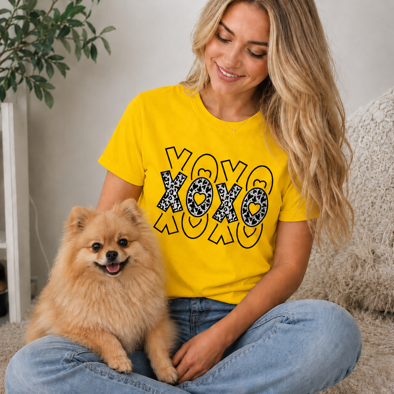 Woman wearing a yellow t-shirt with 'XOXO' design sitting with a small dog indoors.