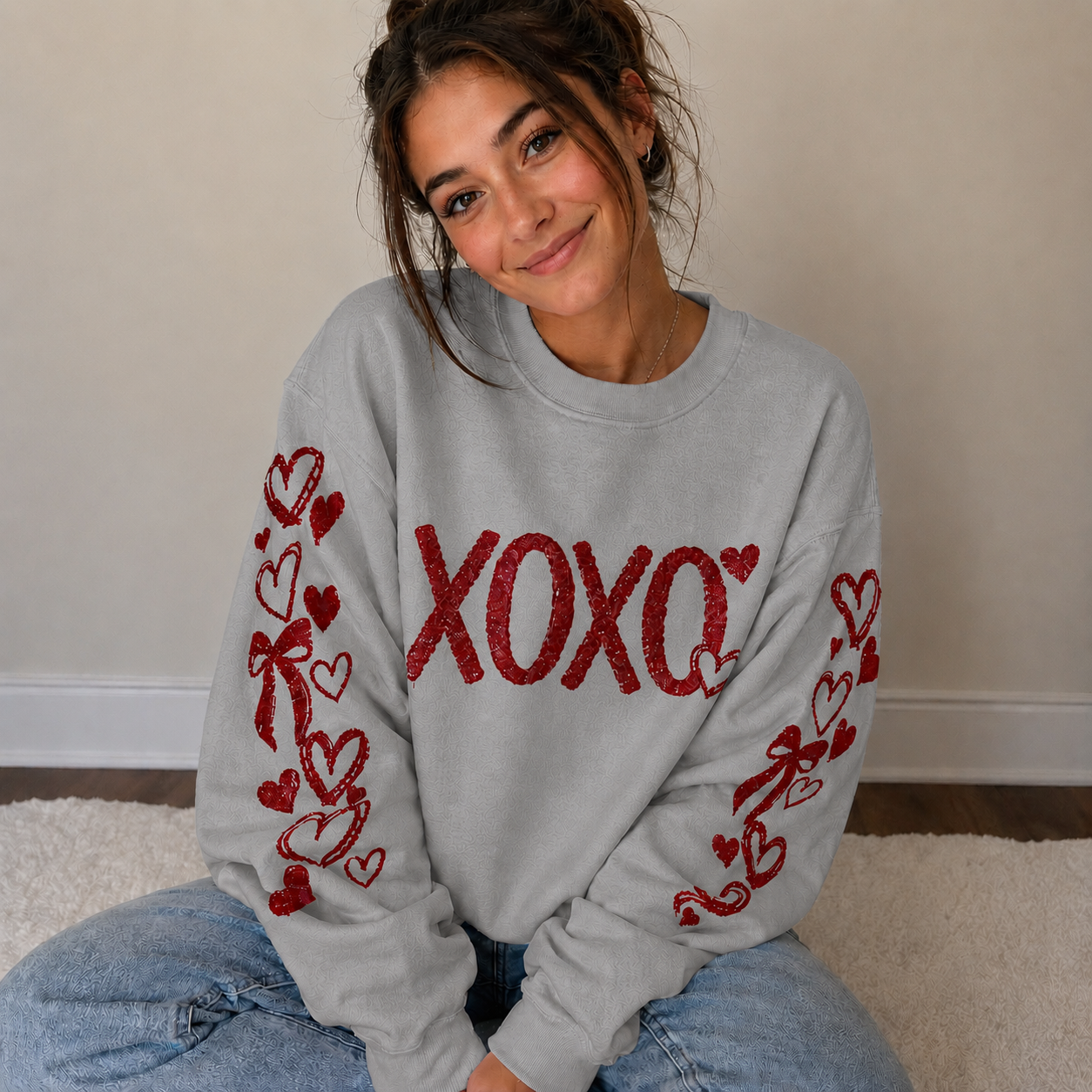 Woman wearing a gray sweatshirt with red 'XOXO' and heart designs, sitting on a carpeted floor.