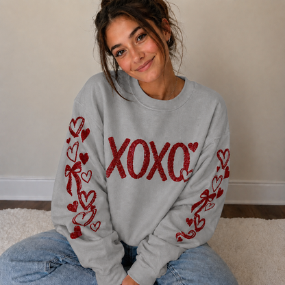Woman wearing a gray sweatshirt with red 'XOXO' and heart designs, sitting on a carpeted floor.