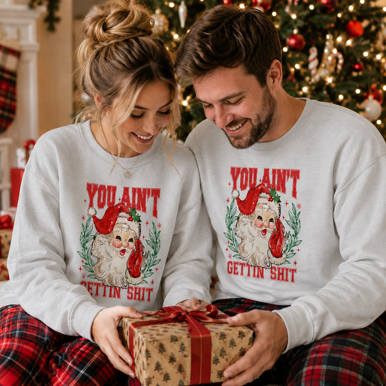 A man and woman wearing matching white crewneck sweatshirts with a Christmas graphic of a Santa dog and the text 'YOU AIN'T GETTIN' SHIT' sit together holding a wrapped gift box in front of a decorated Christmas tree.