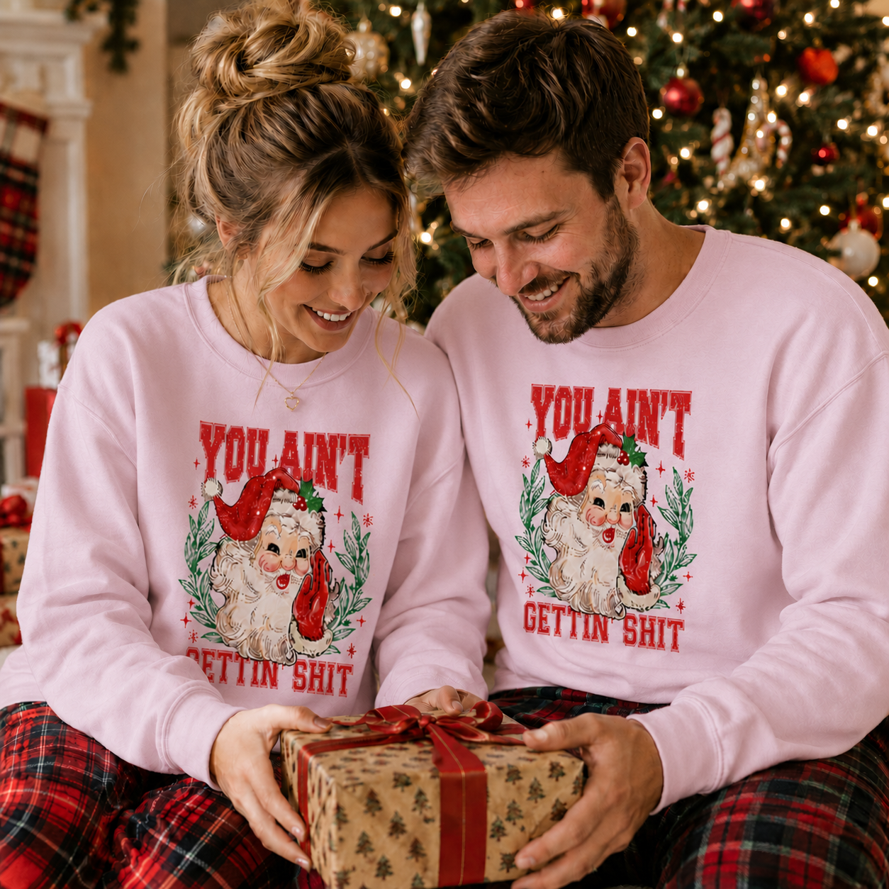 A man and a woman sitting together in a cozy holiday setting, wearing matching light pink crewneck sweatshirts with a vintage-style dog wearing a Santa hat graphic and the text 'YOU AIN'T GETTIN' SHIT'. They are holding a wrapped gift box with a red ribbon.
