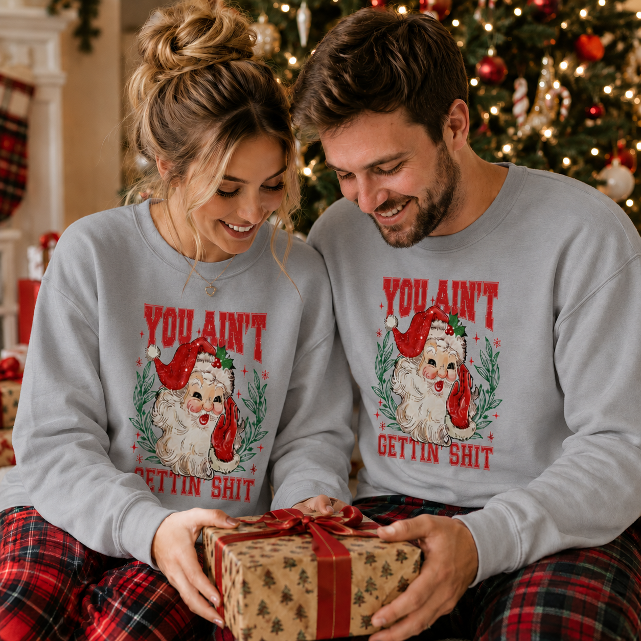 A couple wearing matching light gray Christmas sweatshirts with a dog and Santa graphic and red text, holding a wrapped gift box in a decorated holiday home setting.