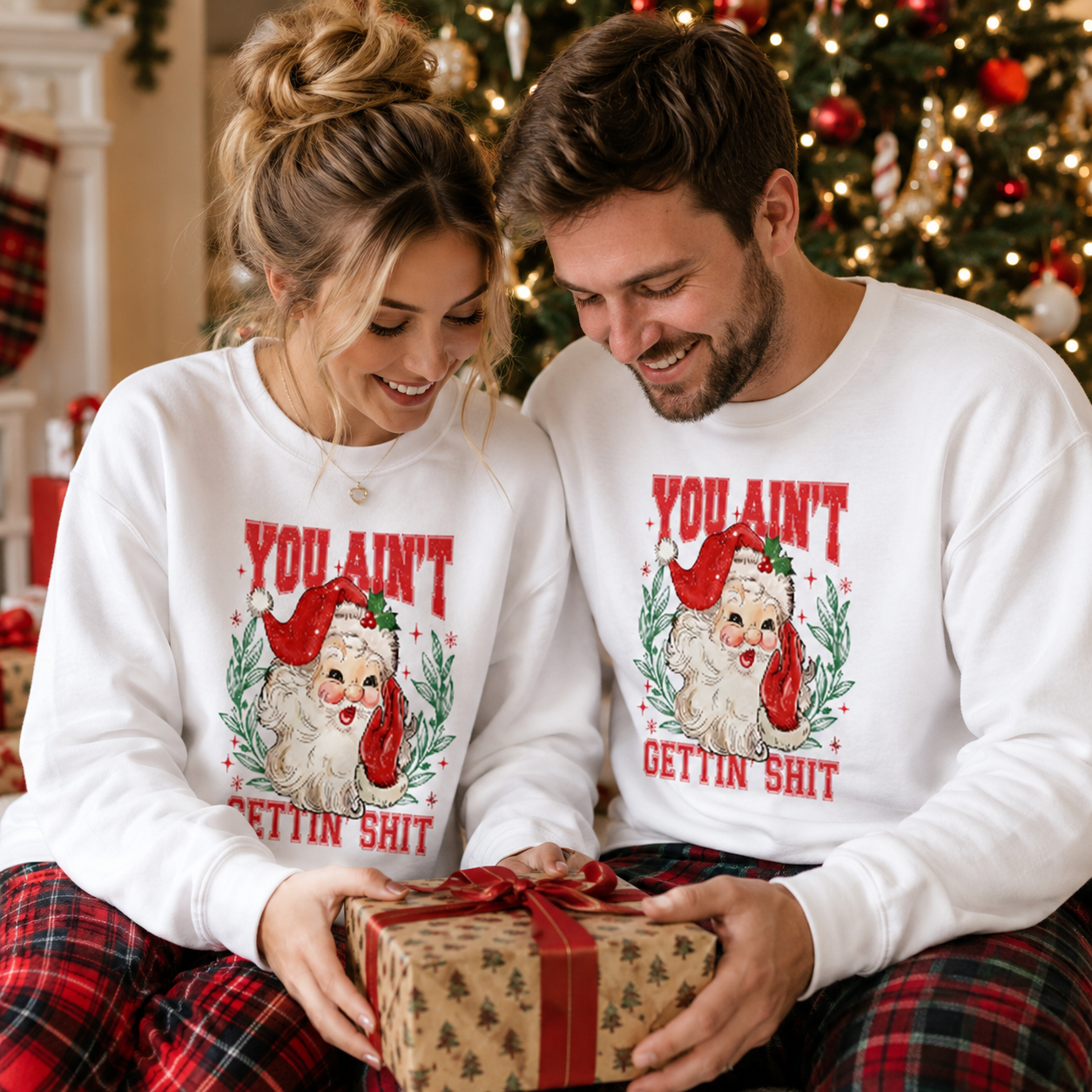 A man and a woman wearing matching white Christmas sweatshirts with a Santa graphic and the text 'YOU AIN'T GETtin' SHIT' are sitting together in front of a lit Christmas tree, holding a wrapped gift.